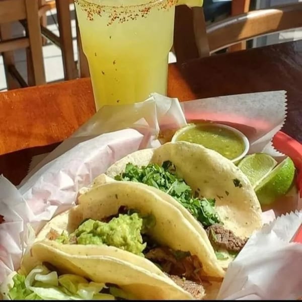 Three tacos with meat, guacamole, and cilantro, served with lime, salsa, and a drink.