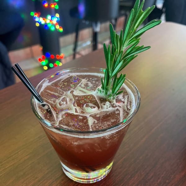 A dark red cocktail with ice, a rosemary sprig, and a black straw on a wooden table.