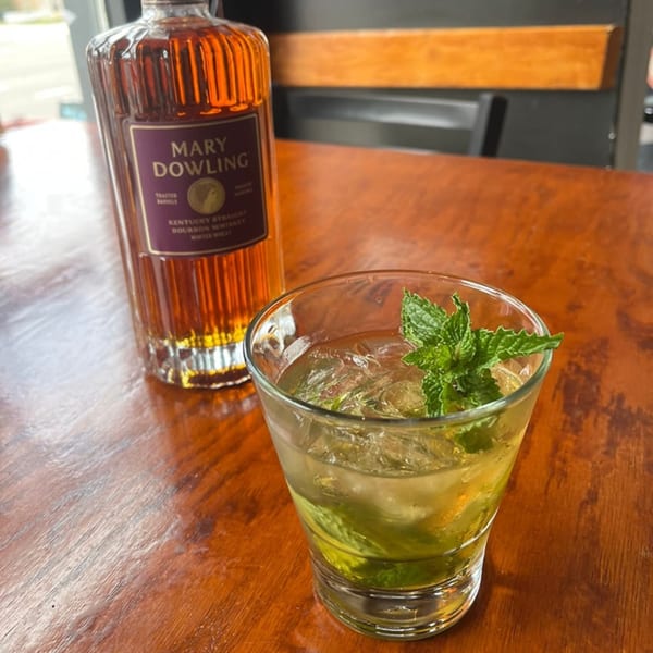 A glass of iced bourbon cocktail with mint sits next to a bottle of Mary Dowling bourbon.