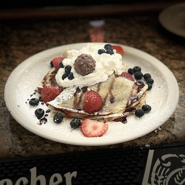 Crepes topped with whipped cream, chocolate, strawberries, blueberries, and powdered sugar on a white plate.