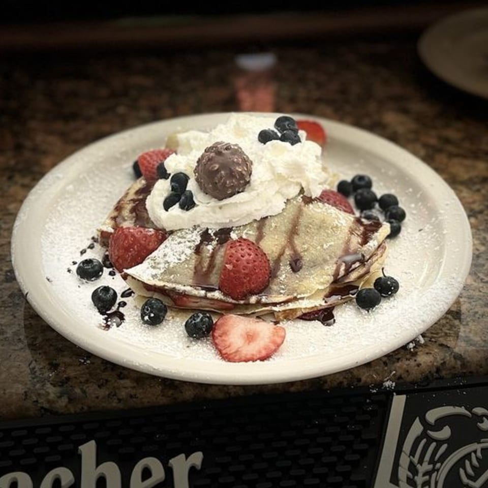Crepes topped with whipped cream, chocolate, strawberries, blueberries, and powdered sugar on a white plate.